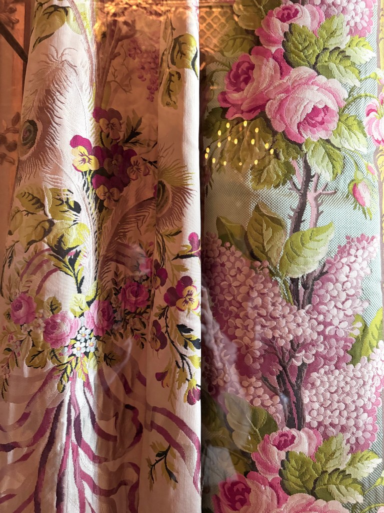 Close-up of luxurious floral fabric featuring large pink roses, wisteria clusters, pansies, and botanical motifs in shades of pink, mauve, green, and gold on a cream background — elegant and richly textured.