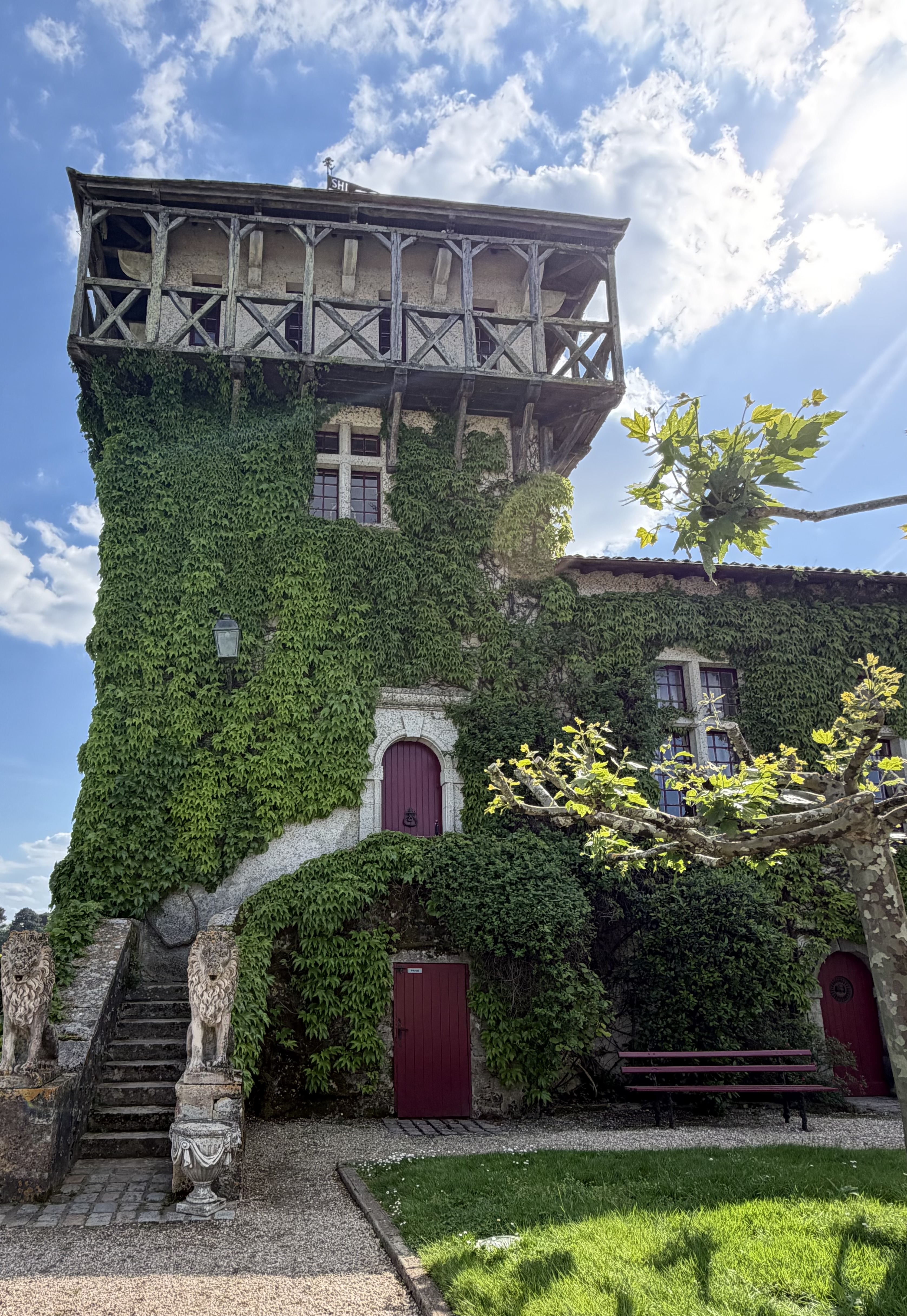 Exterior of a historic stone manor or château covered in dense green ivy, with a rustic wooden balcony on the upper level, arched burgundy doorways, ornate stone urns, and lush surrounding greenery under a bright blue sky.