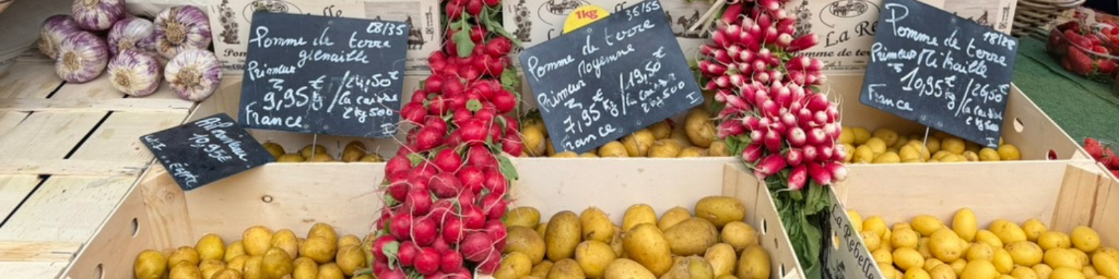 Wooden crates of French primeur potatoes and bundles of fresh radishes with handwritten chalkboard price signs at an outdoor French market